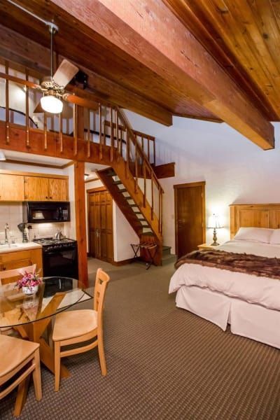 Cozy loft apartment with a bed, kitchen, dining area, and wooden stairs leading to a second level. Warm colors and exposed beams create a rustic atmosphere.