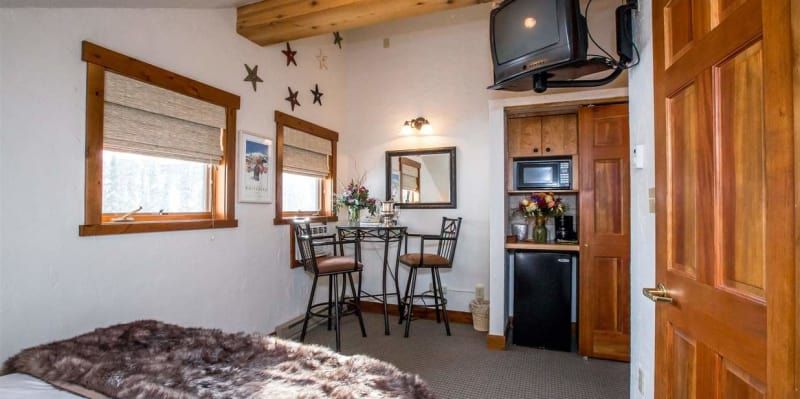 A small hotel room with a bed, table, windows, and a small kitchenette. The walls are white, and the wood trim and door are a warm brown.