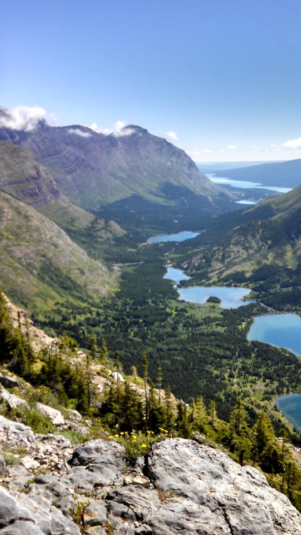 A mountainous landscape with several blue lakes and lush green forests. A clear, sunny sky tops the scene.
