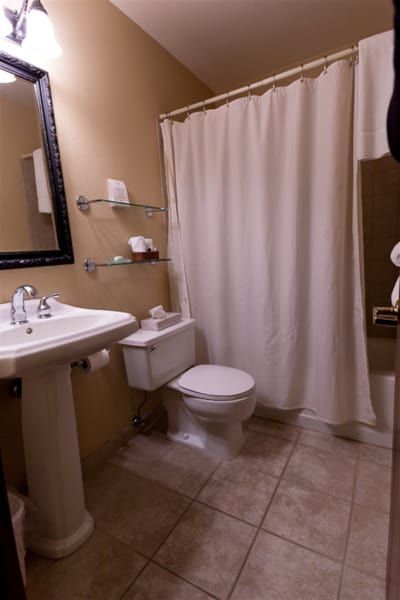 Bathroom with a white pedestal sink, toilet, and shower/tub. Tan walls, beige tile flooring, and a white shower curtain.