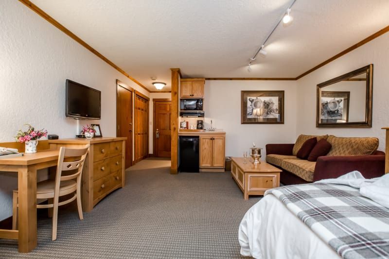 A hotel room with a bed, sofa, desk, and kitchenette. Neutral tones with wood accents and artwork.