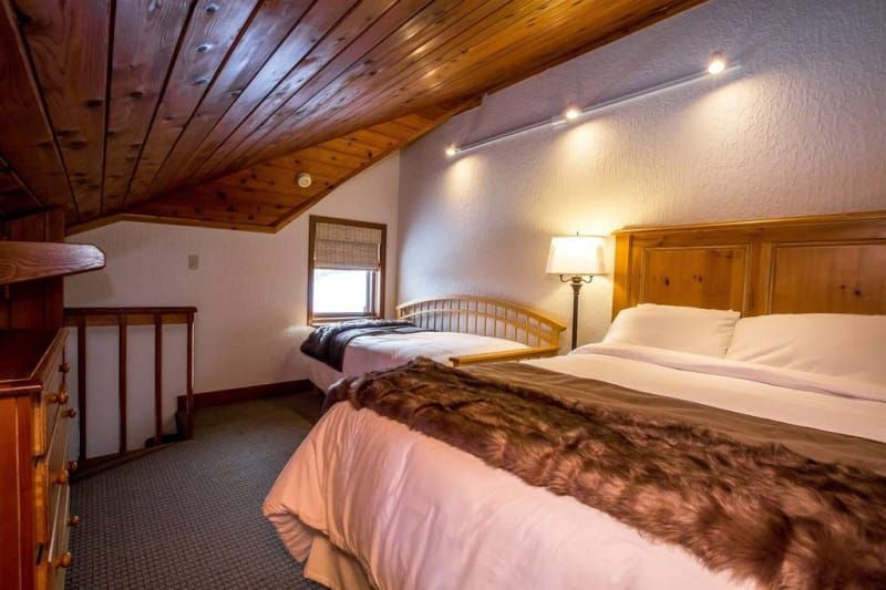 Cozy bedroom with wooden ceiling and furniture. A queen bed has a faux fur throw, and a twin bed sits near a window.