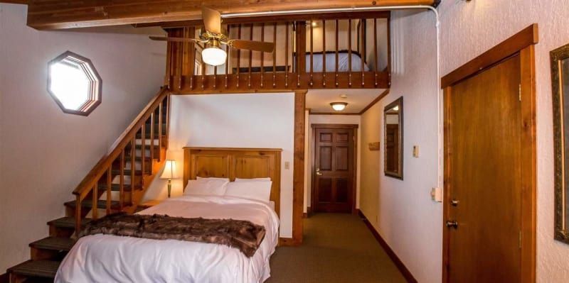A bedroom with a loft and wooden accents. A bed is under the loft, with a brown comforter. A hallway leads to a wooden door.