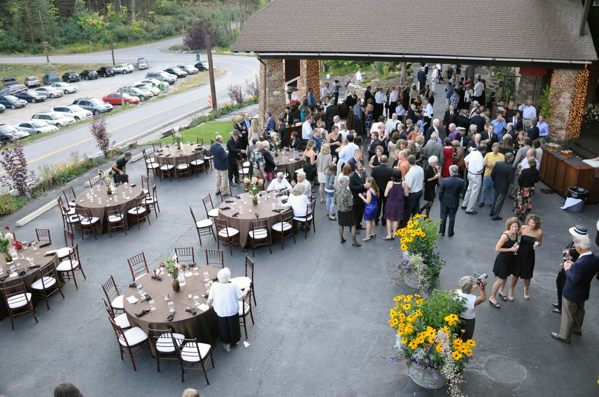 An outdoor event with guests mingling. Tables are set with round brown tablecloths and striped chairs. A parking lot and trees are in the background.