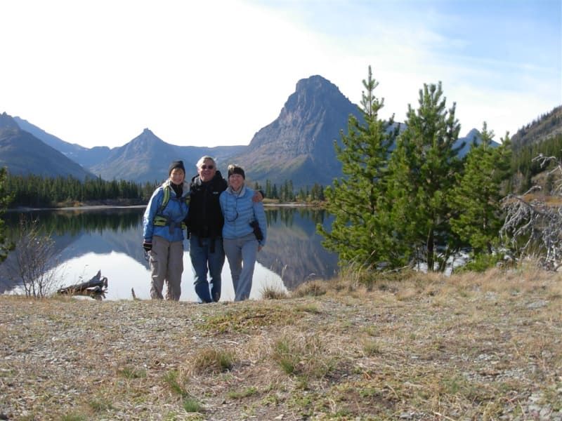 Three people stand by a calm lake with mountains in the background. Blue sky, sunny day.