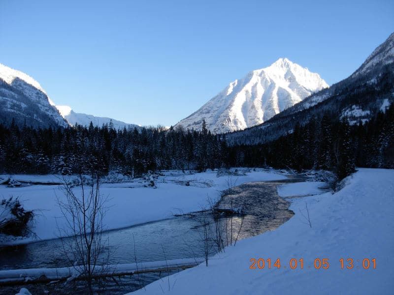 Snow-covered mountains frame a winding river; forest and snow cover the foreground on a clear, sunny day.