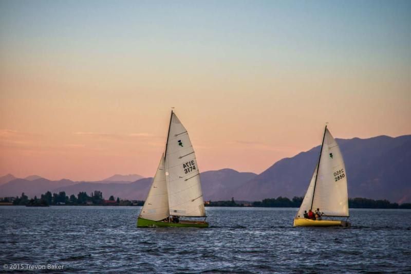 Two sailboats with raised white sails on a lake at dusk, mountains in the background, under an orange and blue sky.