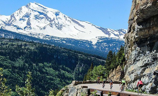 Cyclists riding along a mountain road with a snow-capped mountain backdrop. Green trees and blue sky.