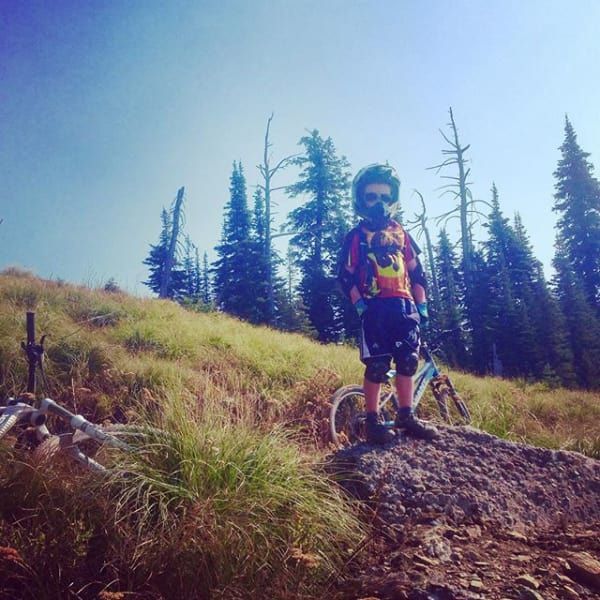 A young mountain biker in protective gear stands on a rock with their bike in a sunny mountain setting.