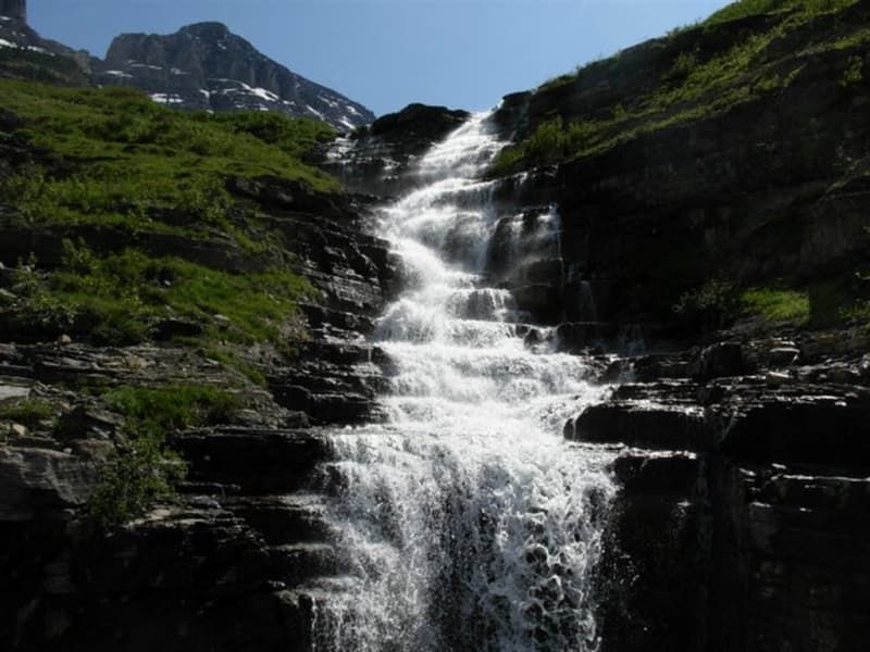 Waterfall cascading down a rocky mountainside, surrounded by green vegetation, under a sunny sky.