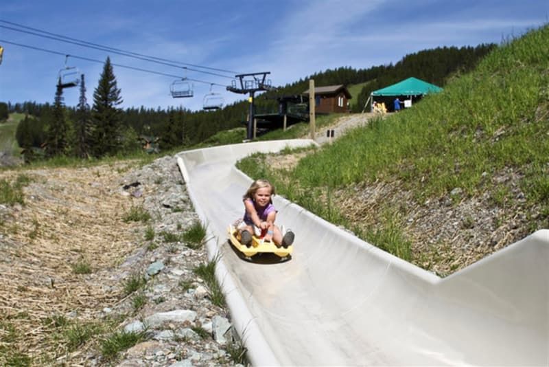 Girl on a yellow sled going down a concrete slide in a mountain setting. Green grass and ski lift in the background.