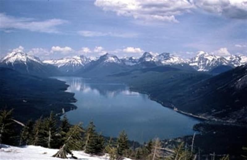 Lake surrounded by mountains, snow-capped peaks reflecting in the blue water. Trees in the foreground.