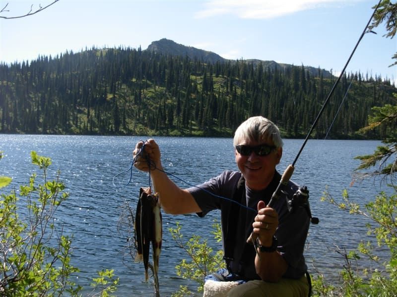 Man fishing at a lake, holding up two fish. He smiles, with trees and a mountain in the background.