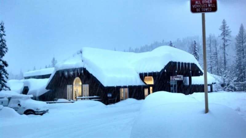 Snow-covered building with lights on, snow-covered ground and trees. A signpost on the right.