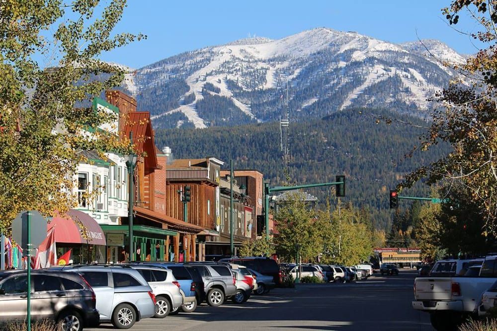 Street scene with parked cars, shops, and a snow-capped mountain in the background. Bright blue sky.