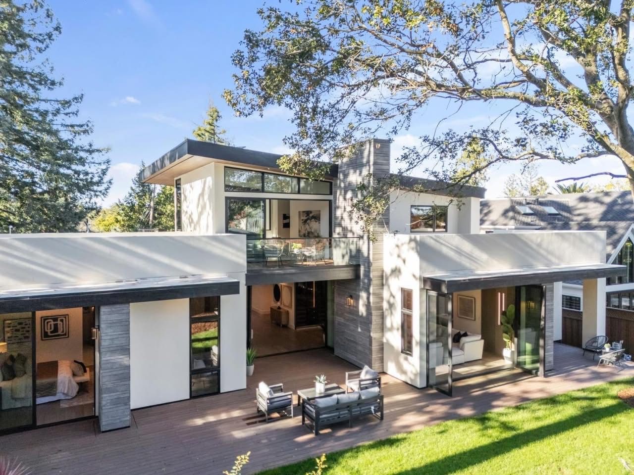 Modern two-story house with large windows, patio seating, and trees in the front yard