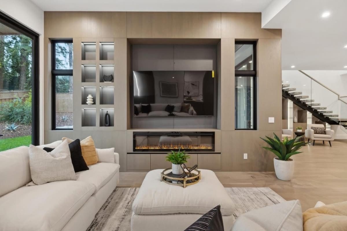 Modern living room with beige sectional, built-in fireplace, wall TV, and staircase in the background