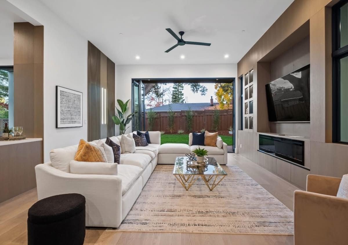 Modern living room with white sofa, fireplace, wall TV, and large glass doors to a backyard.