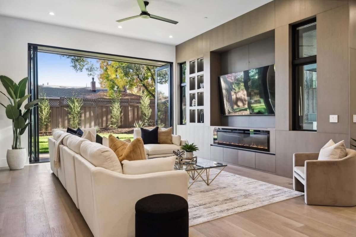 Modern living room with beige sofa, wall-mounted TV, indoor plants, and open glass doors to a patio.