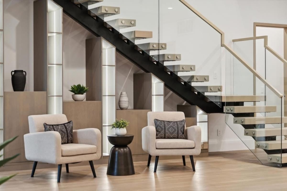 Modern living room with two beige chairs, black side table, staircase, and light wood floors.