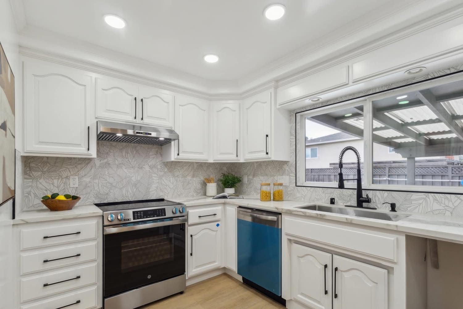 Bright white kitchen with stainless stove, large window sink, and blue dishwasher panel