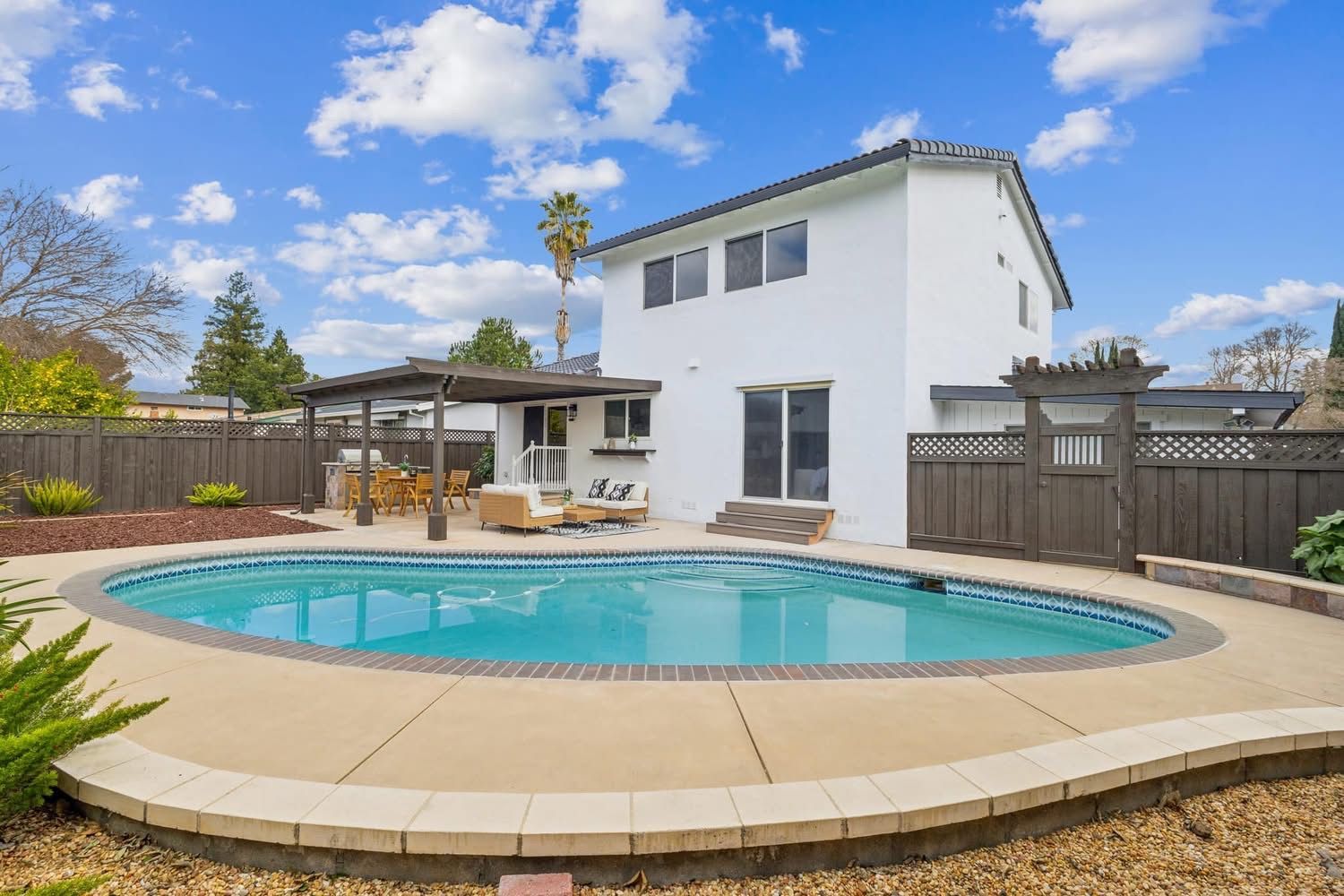 Backyard with a round swimming pool, white two-story house, patio, and wooden fence under a blue sky