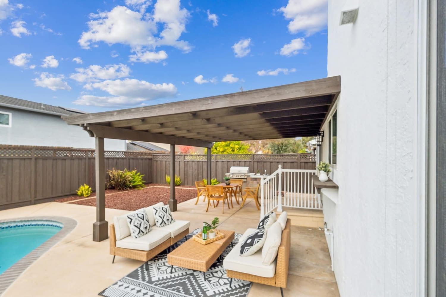 Covered patio seating beside a pool, with a wooden pergola, outdoor sofa, and blue sky overhead