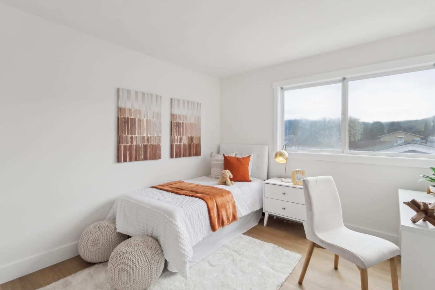 Minimal white bedroom with a single bed, desk, chair, wall art, and large window.