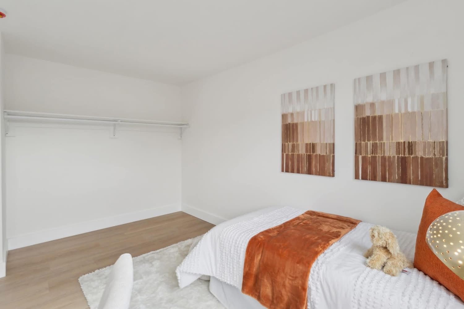 Minimal bedroom with white walls, wood floor, bed with orange throw, and two abstract wall artworks.