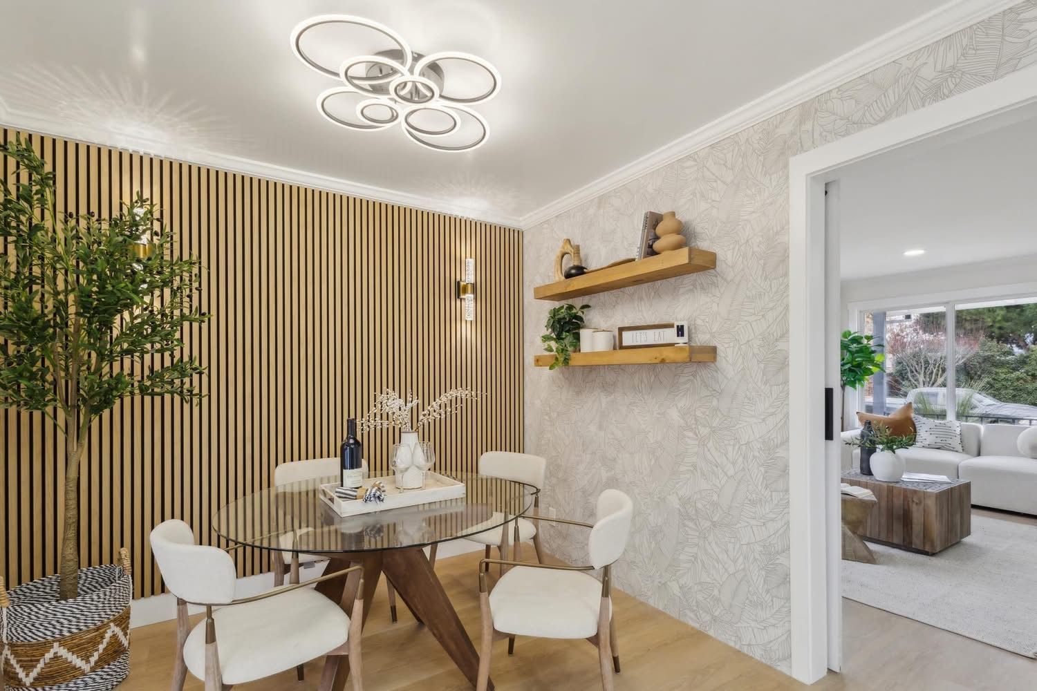 Bright dining room with round table, white chairs, wood slat accent wall, and doorway to a sunlit living area