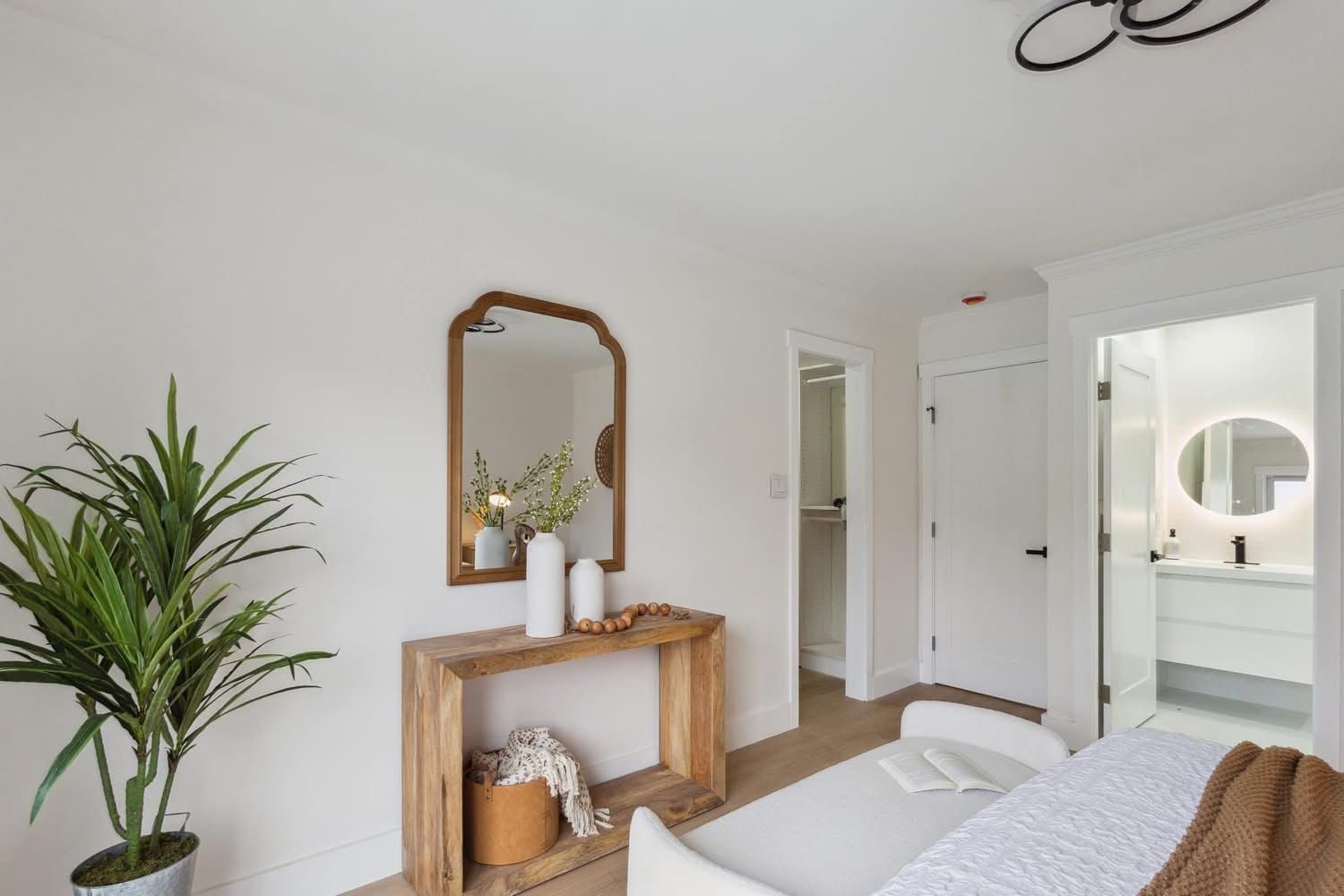 Bright minimalist bedroom with wood console, mirror, plant, and open doors to a bathroom.