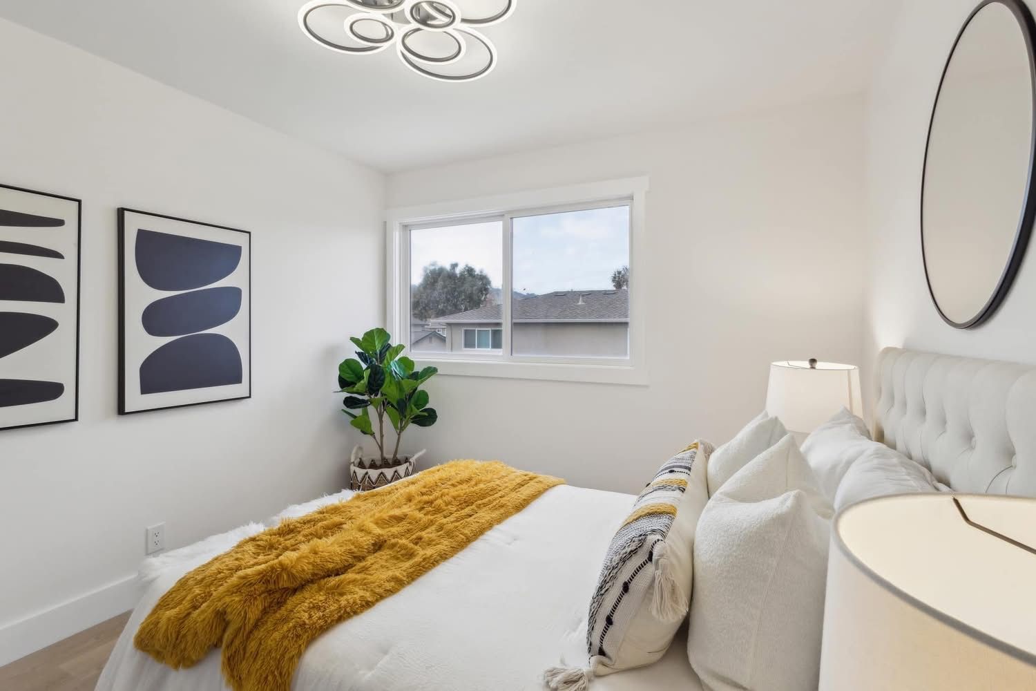 Bright modern bedroom with white walls, mustard throw blanket, framed art, plant, and a window view