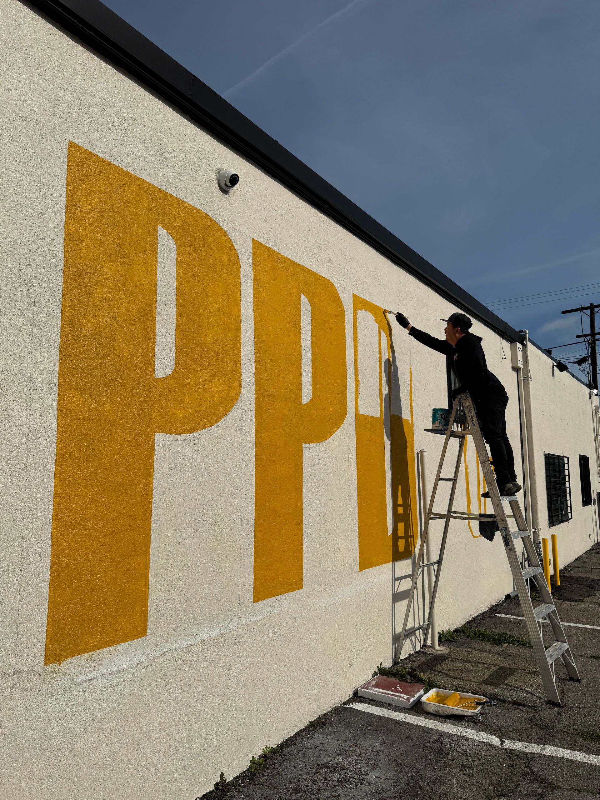 Worker on ladder painting large yellow letters on a white wall outdoors