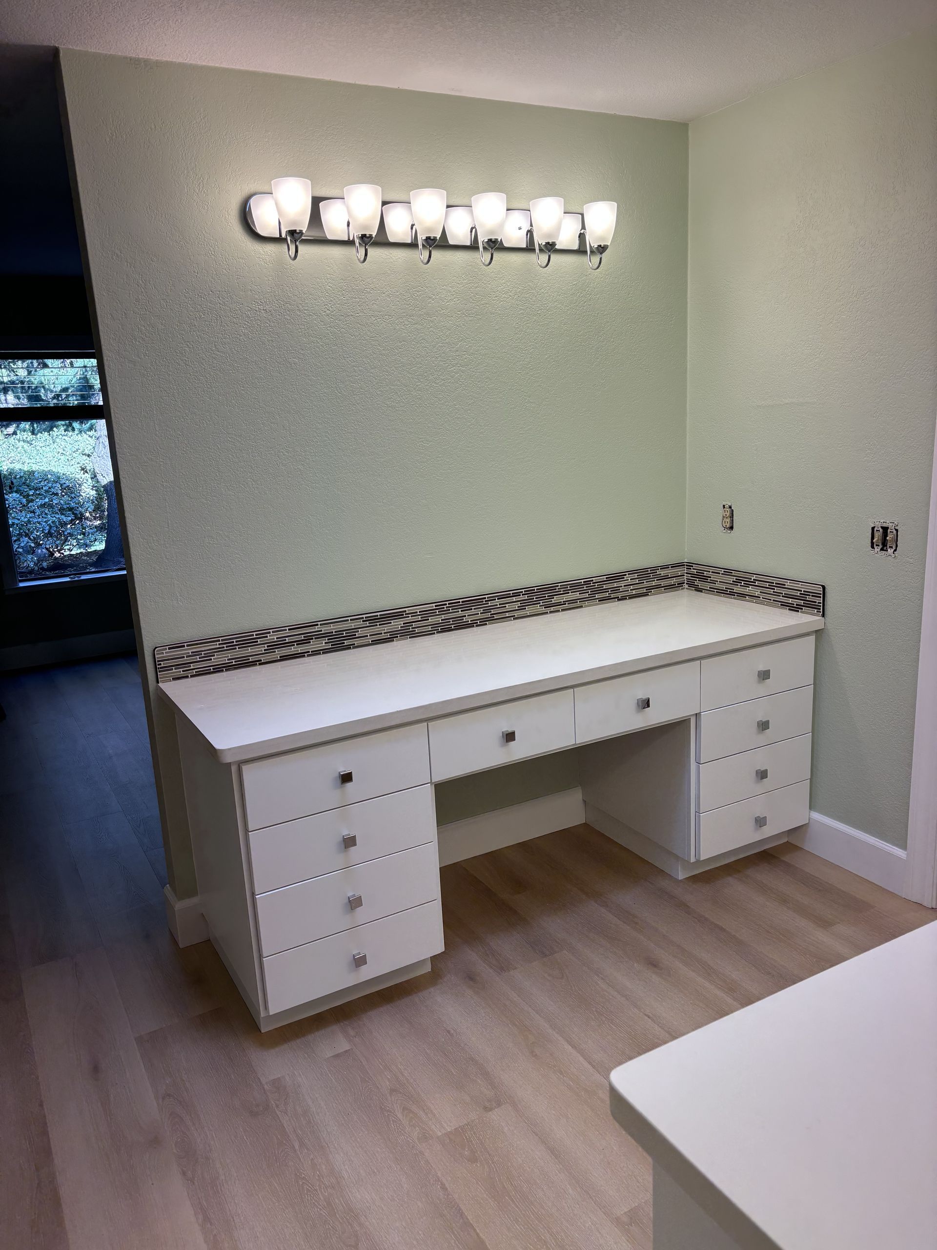 Built-in white desk with drawers in a light green room, beneath a wall sconce and stone backsplash