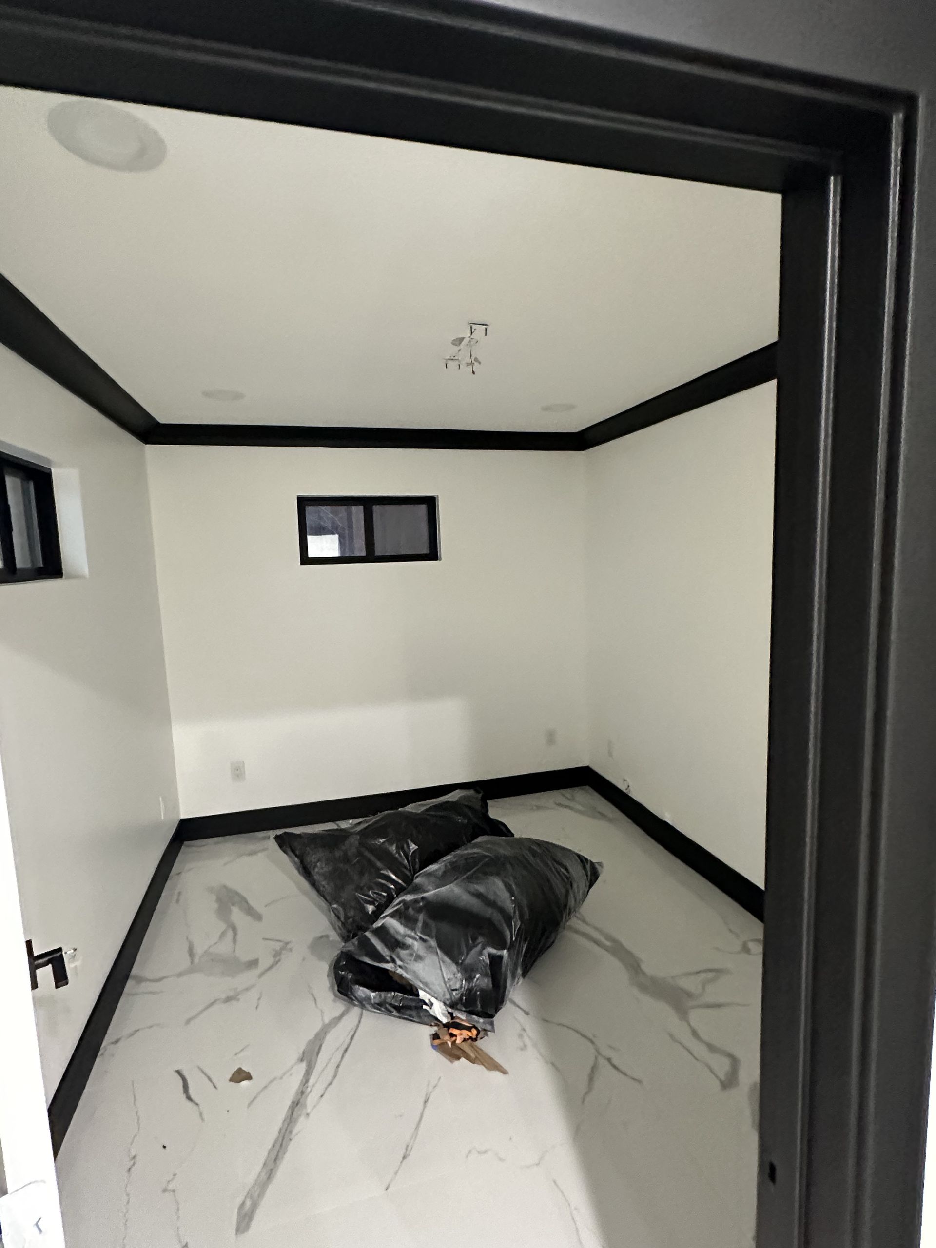 Small empty room with white walls, black trim, and a black plastic-covered bundle on the floor.