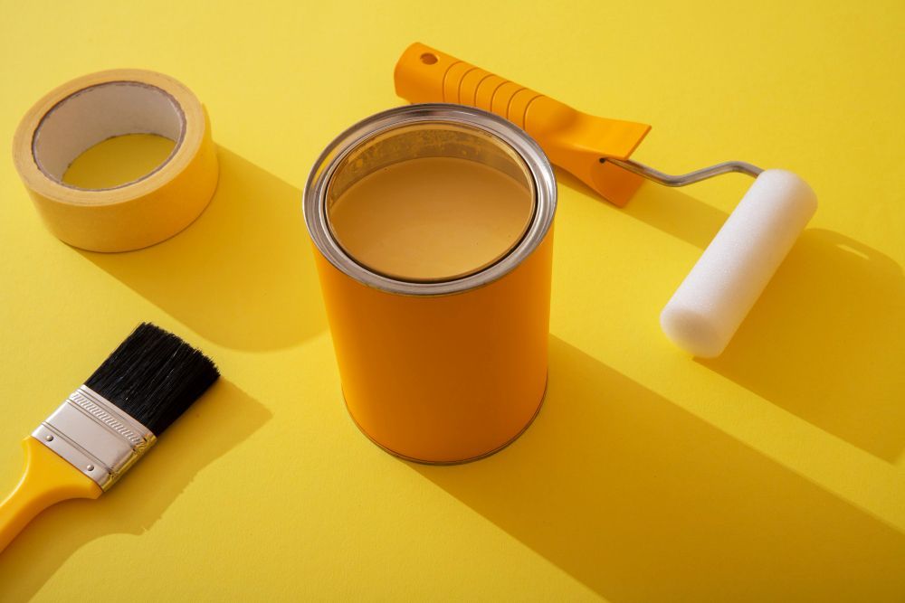 A can of yellow paint , a brush , a roller and tape on a yellow background.