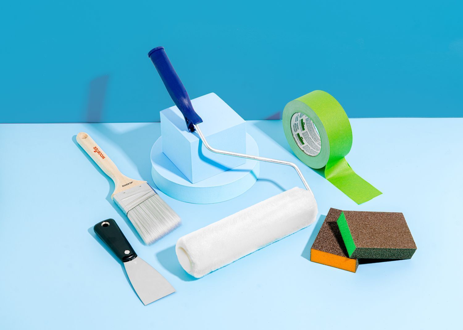 Painting tools on a light blue surface, including brushes, tape, a roller, and a paint tray