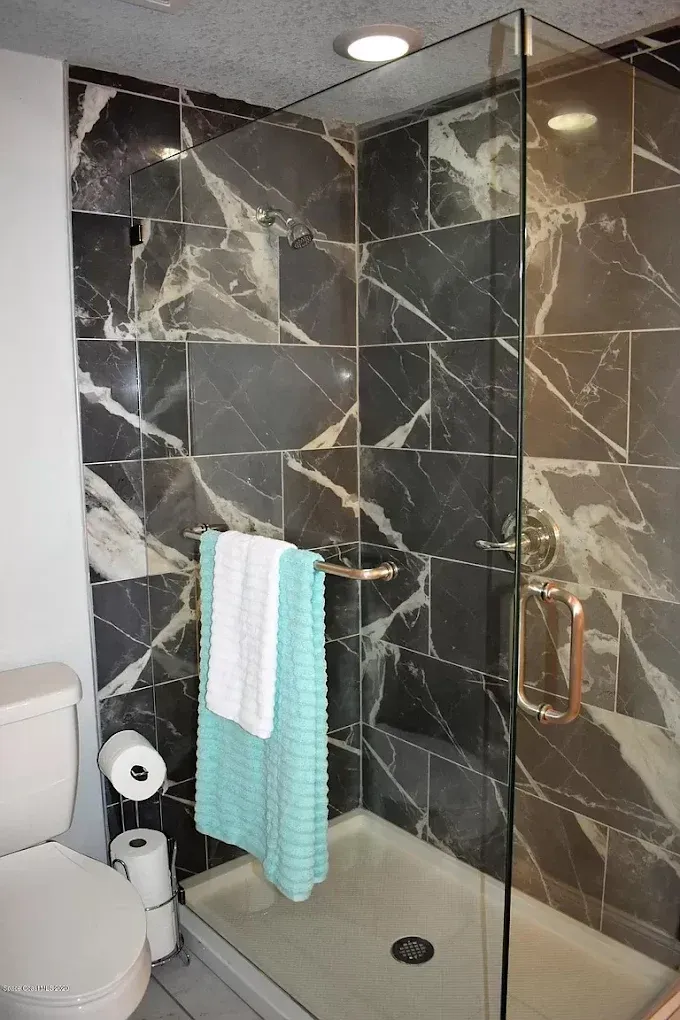 Bathroom with dark marble tile shower, glass door, white toilet, and towels.