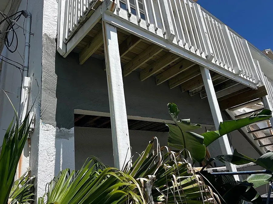 Exterior of a building with a white balcony and wooden deck. Plants in the foreground, blue sky.