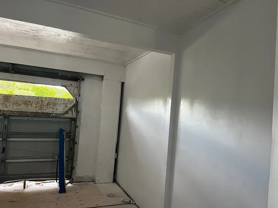 Interior view of a room with a white painted wall, doorway to garage. Daylight streams through the opening.