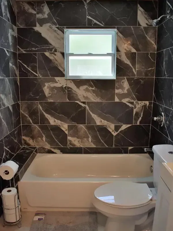 Bathroom with white tub and toilet, dark marbled tile walls, small window.