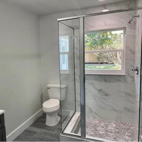 Bathroom with a glass shower, toilet, and window with natural light. Gray and white color scheme.