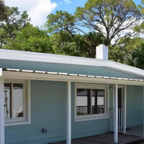 Blue-sided house with white trim, windows, and porch, set against green trees and a cloudy sky.