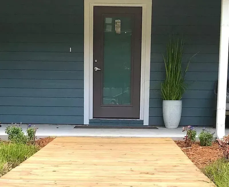 Wooden walkway to a teal house with a brown door, potted plant, and small landscaping.