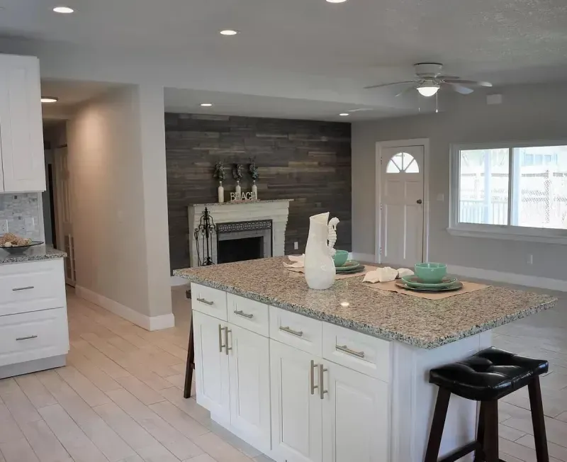 Modern open-concept kitchen with white cabinets, island, granite countertop, and fireplace with wood paneling.