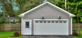 Gray garage with a white door and garage door; greenery in the background.