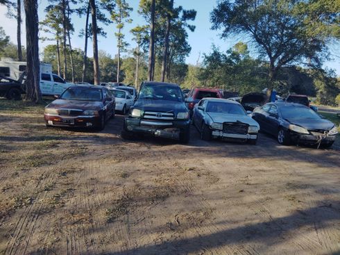 Cars parked on a dirt lot surrounded by trees. The vehicles appear older, some damaged.