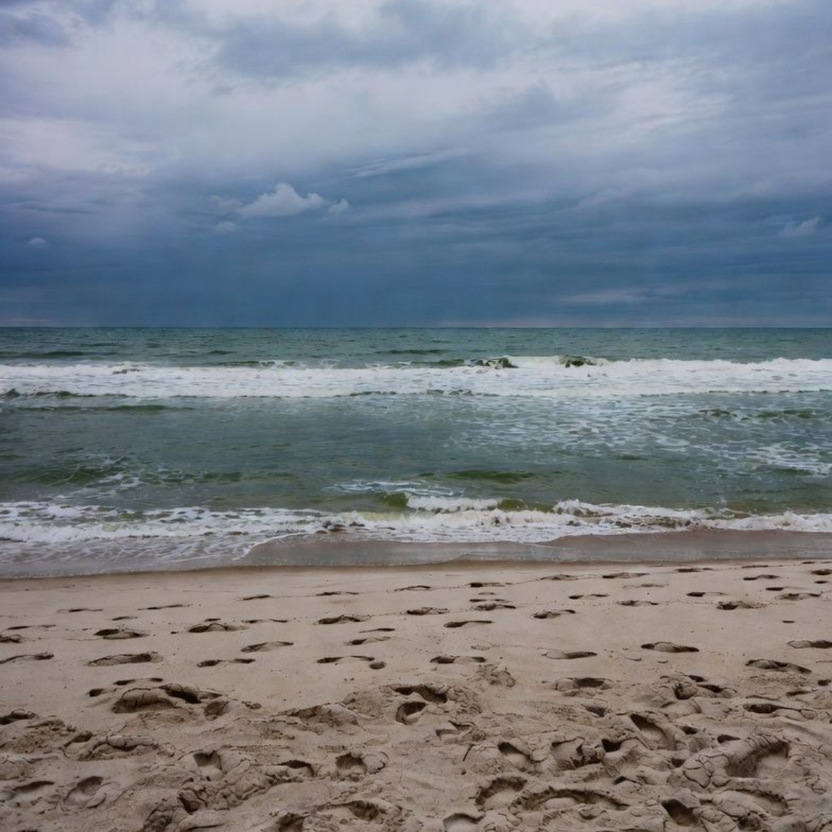 Sandy beach with footprints, waves, and cloudy sky.