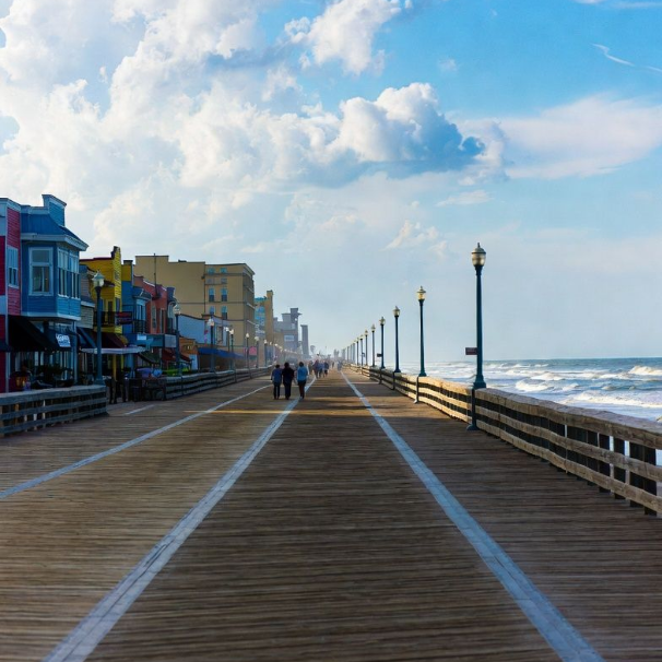 Wooden boardwalk along colorful buildings, people walking towards the ocean under a cloudy sky.