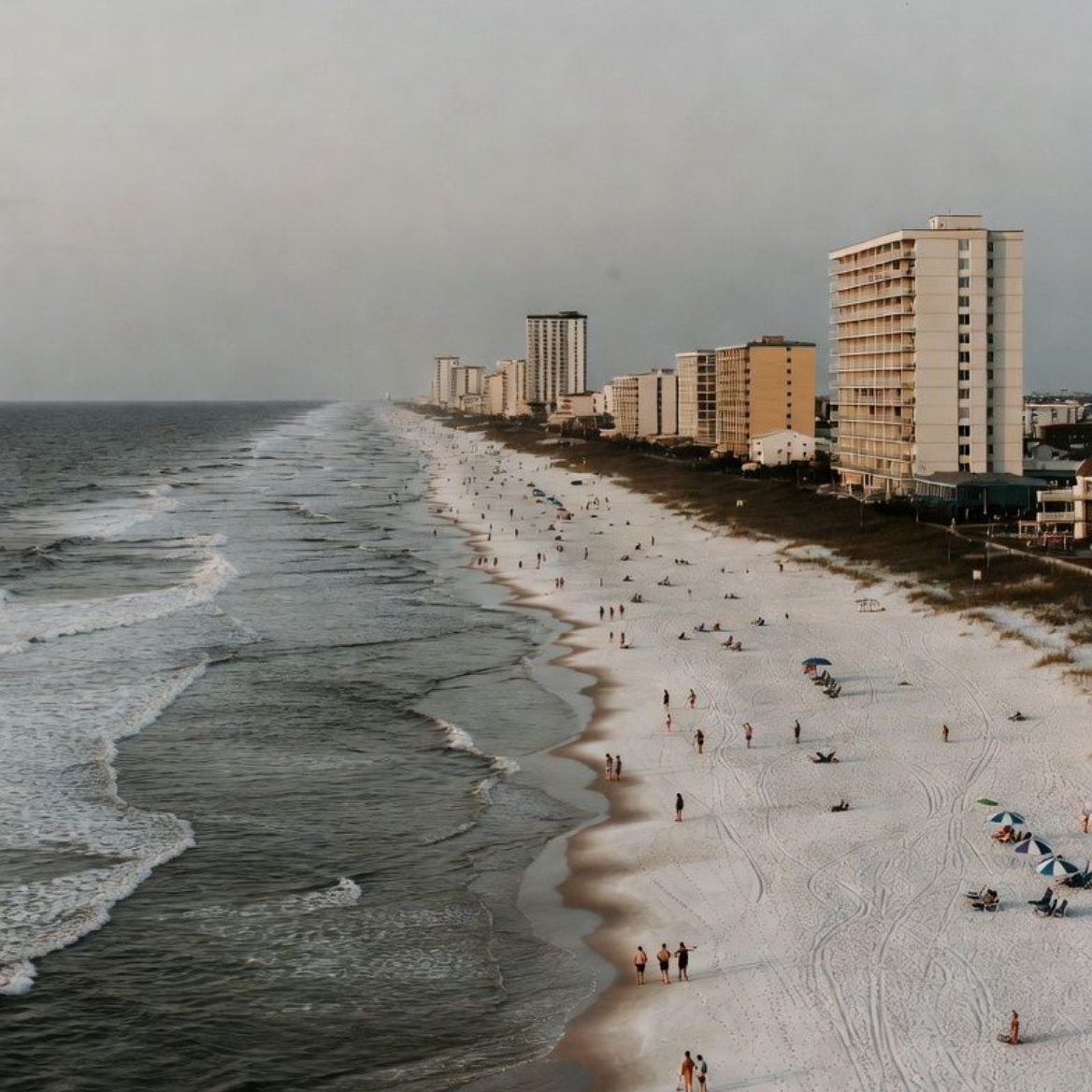Beach with white sand, waves, and buildings along the shore. People are scattered on the beach.